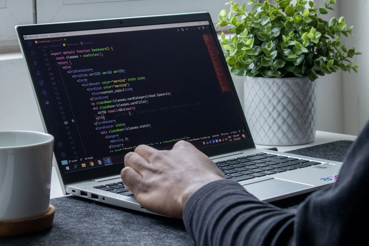 A person typing code on a laptop beside a plant and mug.